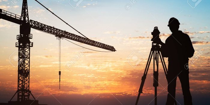 Silhouette Of A Surveyor Standing With Equipment Near Crane At Construction Site During Sunset